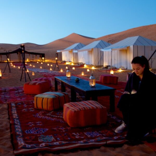 A tourist sitting in a desert camp in Merzouga with tents and candles in the back.