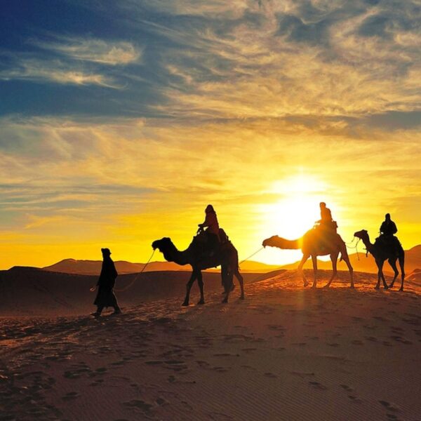 3 personas montando en camello durante la puesta de sol con nuestro viaje de 4 días por el desierto de Errachidia a Marrakech.